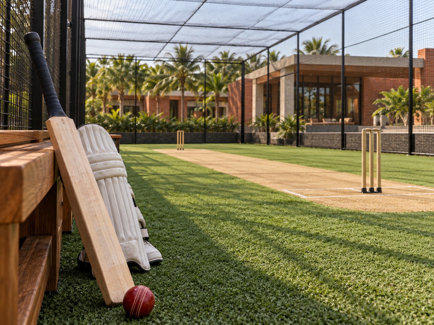 close sideline view of a wooden cricket bat, red cricket ball and batting pads beside the turf, cricket pitch and black enclosure nets visible beyond